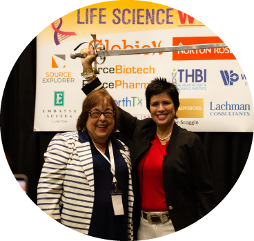 A circular image of two women standing on stage, smiling. The one on the right is raising a broadsword with an orange-tinted sponsor banner backdrop that reads, The Life Science Women's Conference.